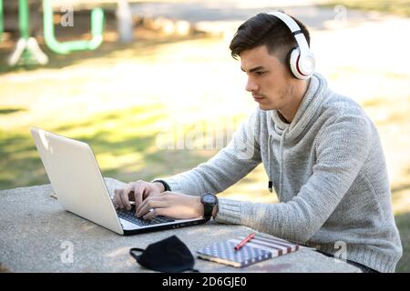 Giovane uomo che studia con il computer mentre ascolta la musica con cuffie nel parco senza maschera Foto Stock