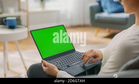 Giovane donna a casa seduta su un lettino funziona su un computer portatile con schermo verde mock-up. Ragazza che usa il computer, navigare attraverso Internet, guardare Foto Stock