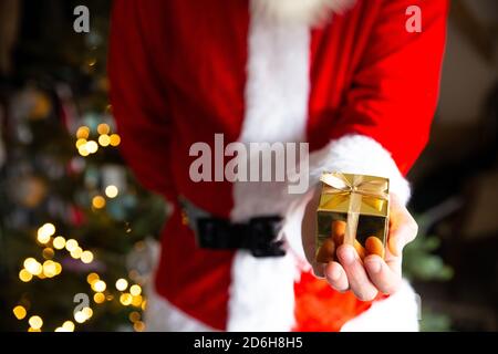 Uomo in costume di Babbo Natale vicino albero di Natale che tiene una scatola di regalo dorata, sfondo del presente di Natale con le luci del bokeh Foto Stock