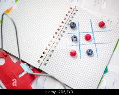 Concetto di ritorno a scuola. TIC-tac-TOE gioco di lamponi e mirtilli su un copybook con il libro titolare in primo piano. Studiare con divertimento. Foto Stock