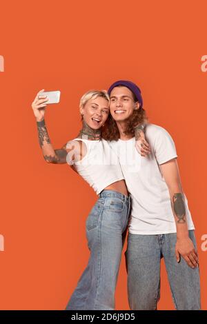Stile libero. Giovane coppia tatuata in piedi isolati su orange hugging prendendo selfie sullo smartphone in posa per la fotocamera divertente Foto Stock