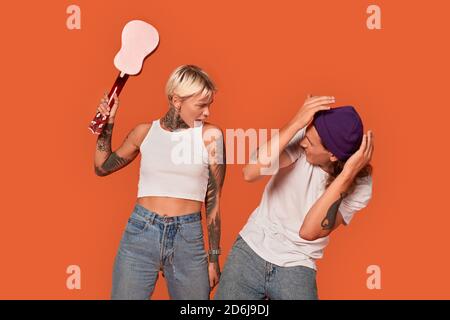Stile libero. Giovane coppia tatuata in piedi isolata su fidanzata arancione battendo il ragazzo con ukulele gridando arrabbiato Foto Stock