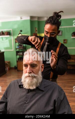 giovane barbiere e dreadlock afro-americani, con maschera di prevenzione pandemica del coronavirus, combe con un pettine e le sue mani un uomo caucasico bianco maturo Foto Stock