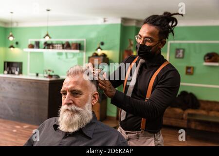 giovane barbiere e dreadlock afro-americani, con maschera di prevenzione pandemica del coronavirus, combe con un pettine e le sue mani un uomo caucasico bianco maturo Foto Stock