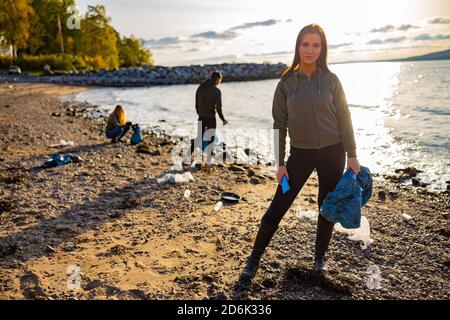 Donna giovane seria che pulisce la spiaggia per la plastica con i volontari durante tramonto Foto Stock