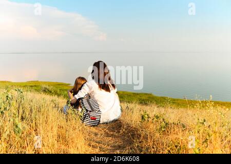 Vista posteriore di donna irriconoscibile abbracciando la ragazza mentre si siede sopra erba secca e ammirando il pittoresco paesaggio marino durante il fine settimana estivo Foto Stock