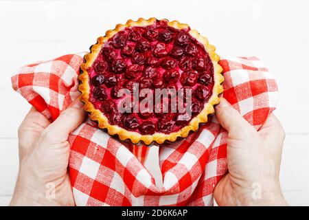 Le mani dello chef tengono un tartlet appena sfornato con ciliegia in un asciugamano rosso e bianco. Foto Stock