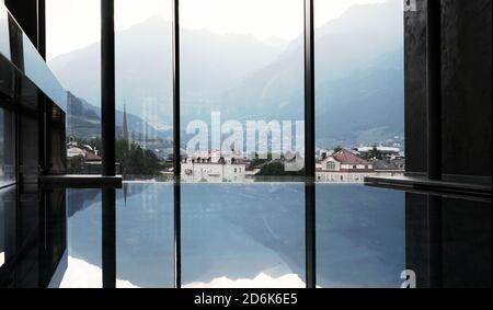 Vista degli edifici di Merano, del Tirolo del Sud e delle Alpi Italiane, delle montagne dall'interno di una lussuosa piscina interna. Foto Stock