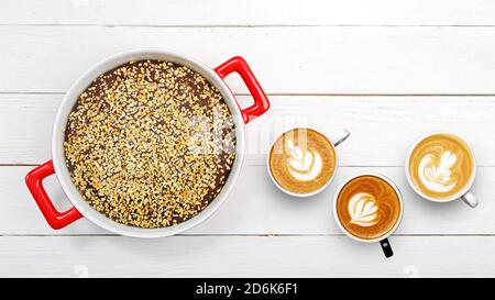 Brownie fatte in casa cosparse di briciole di noci in una forma di ceramica bianca e rossa e tre tazze di caffè su un tavolo di legno bianco. Vista dall'alto. Foto Stock