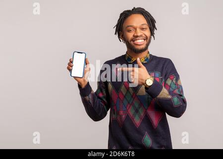 Felice uomo africano soddisfatto con Dreadlock e barba che mostra smartphone con vuoto bianco sinceramente sorridente, puntando sulla pubblicità. Monolocale interno Foto Stock