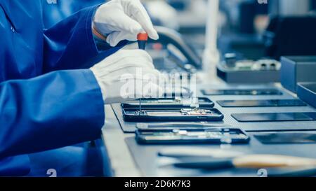 Primo piano di una lavoratrice femminile di una fabbrica di elettronica in un camice da lavoro blu e occhiali protettivi assemblaggio di smartphone con cacciavite. High Tech Factory Foto Stock