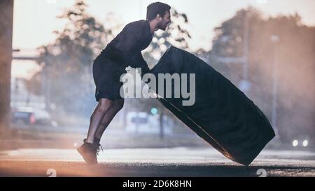 Forte Muscular Fit Young Man sta facendo esercizi in strada. Sta girando un pneumatico pesante grande in un ambiente urbano. Foto Stock