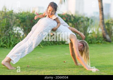 Giocosa bambina che si arrampica sulla schiena della mamma durante un divertimento in famiglia al parco durante l'estate. Foto Stock