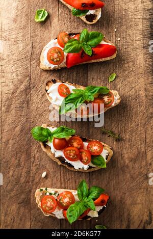 Bruschette con pomodori, formaggio cremoso, paprika alla griglia e basilico su sfondo ligneo, vista dall'alto. Tradizionale spuntino italiano - brusc alla griglia Foto Stock