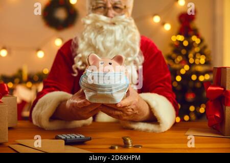 Babbo Natale alla scrivania con soldi e calcolatrice che tiene il piggy banca che indossa una maschera medica Foto Stock