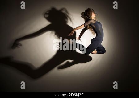 donna al buio esegue il salto ginnico, su uno sfondo bianco c'è un'ombra da una forma. Fotografia da studio. Foto Stock