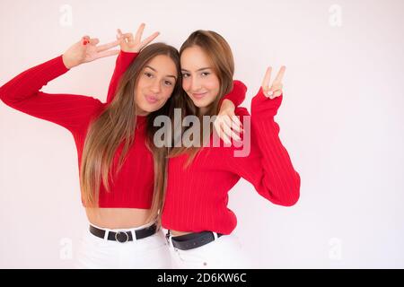 Foto di graziose due fidanzate che mostrano i simboli del segno a V che abbracciano indossando un pullover rosso e jeans isolato sfondo bianco Foto Stock