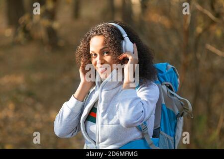 Donna afroamericana sorridente che indossa le cuffie mentre cammini nella foresta con uno zaino. Ascolta la musica e ama la vita. Foto Stock