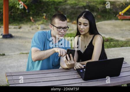 Due giovani studenti seduti nel parco che parlano e studiano sullo smartphone Foto Stock