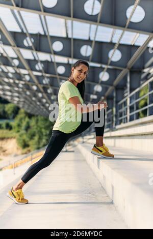 Donna positiva in abbigliamento sportivo che si allunga per le gambe sui gradini Foto Stock