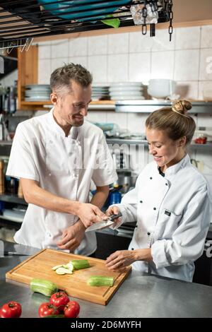 Felice chef professionista che passa coltello affilato al tirocinante affettando fresco zucchine Foto Stock