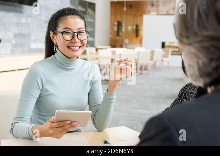 Felice giovane donna d'affari asiatica con tablet seduto di fronte collega maturo Foto Stock