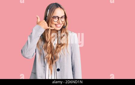 Giovane bella donna bionda che indossa le cuffie dell'agente del call center sorridendo facendo il gesto del telefono con le mani e le dita come parlare al telefono. commun Foto Stock