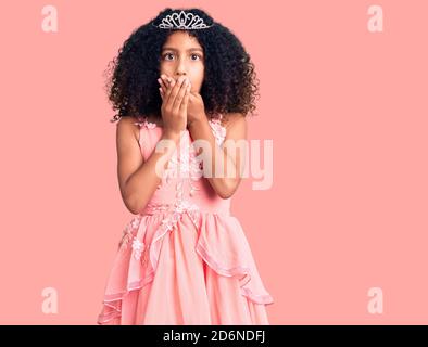 Bambino afroamericano con capelli ricci che indossano la corona della principessa scioccato coprendo la bocca con le mani per errore. Concetto segreto. Foto Stock