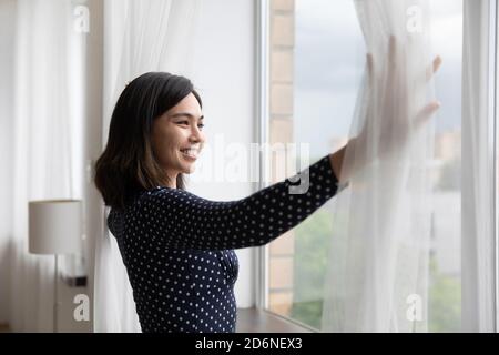 Donna asiatica sorridente apre le tende guardando in lontananza Foto Stock