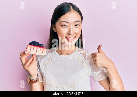 Giovane donna cinese mangiare cheesecake sorridendo felice e positivo, pollice su facendo eccellente e segno di approvazione Foto Stock