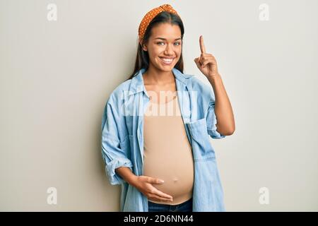 Bella donna ispanica in attesa di un bambino, toccando la pancia incinta sorridendo stupito e sorpreso e puntando con le dita e le braccia sollevate. Foto Stock