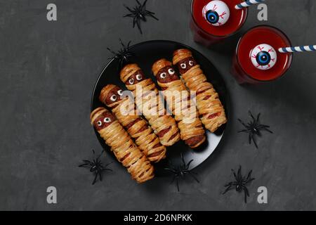 Spooky salsiccia mummie e succo di pomodoro per la festa di Halloween su piatto scuro, vista dall'alto. Disposizione piatta. Foto Stock