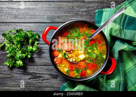 Zuppa di piselli spezzati con salsiccia bavarese, pastinaca, carote con prezzemolo e cipolla verde servita su una pentola rossa antiaderente con un mestolo di zuppa su una da Foto Stock