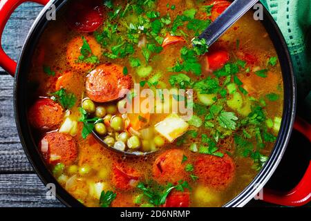 Zuppa di piselli spezzati con salsiccia bavarese, pastinaca, carote con prezzemolo e cipolla verde servita su una pentola rossa antiaderente con un mestolo di zuppa su una da Foto Stock