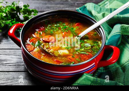 Zuppa di piselli spezzati con salsiccia bavarese, pastinaca, carote con prezzemolo e cipolla verde servita su una pentola rossa antiaderente con un mestolo di zuppa su una da Foto Stock