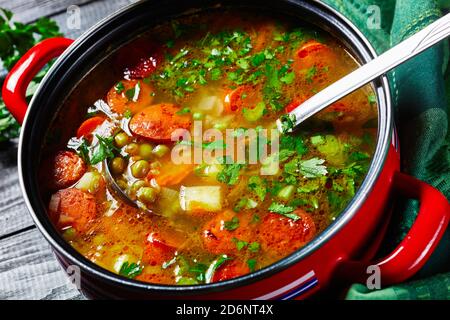 Zuppa di piselli spezzati con salsiccia bavarese, pastinaca, carote con prezzemolo e cipolla verde servita su una pentola rossa antiaderente con un mestolo di zuppa su una da Foto Stock