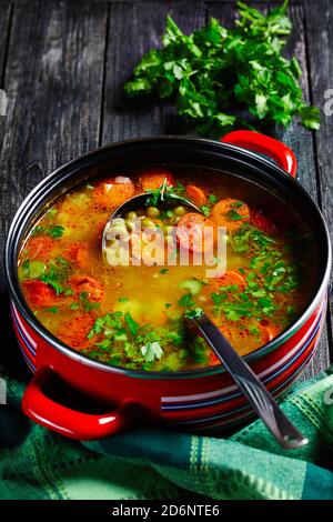 Zuppa di piselli spezzati con salsiccia bavarese, pastinaca, carote con prezzemolo e cipolla verde servita su una pentola rossa antiaderente con un mestolo di zuppa su una da Foto Stock