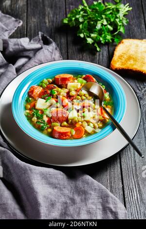 Zuppa tedesca di piselli spezzati con salsiccia bavarese, pastinaca, carote condite con prezzemolo e scalone servita su un piatto blu con un cucchiaio su un bac di legno scuro Foto Stock