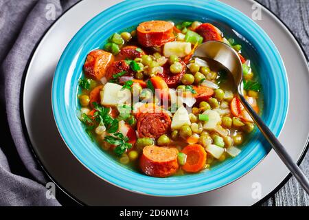 Zuppa tedesca di piselli spezzati con salsiccia bavarese, pastinaca, carote condite con prezzemolo e scalone servita su un piatto blu con un cucchiaio su un bac di legno scuro Foto Stock