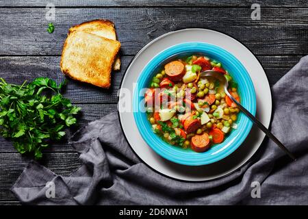 Zuppa tedesca di piselli spezzati con salsiccia bavarese, pastinaca, carote condite con prezzemolo e scalone servita su un piatto blu con un cucchiaio su un bac di legno scuro Foto Stock