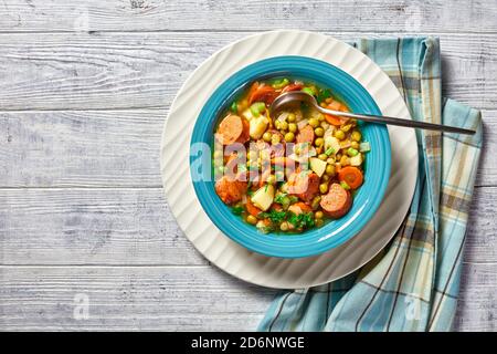 Zuppa tedesca di piselli spezzati con salsiccia bavarese, pastinaca, carote condite con prezzemolo e scalone servita su un piatto blu con un cucchiaio su una zuppa di legno bianca Foto Stock