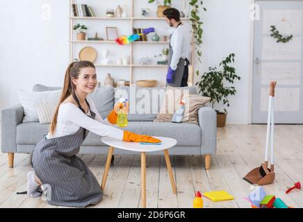 Donna sorridente in grembiule e guanti di gomma con salviette spray polvere da tavolo, marito in mensole Foto Stock