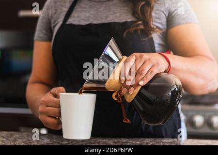 Barista ispanica femminile che versa il caffè in una tazza bianca in una cucina familiare. Donna nera che tiene versa sopra la caffettiera la mattina Foto Stock