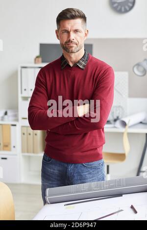 Ritratto verticale di architetto maturo in piedi con braccia incrociate e guardando la macchina fotografica mentre si posa dal tavolo di disegno a. luogo di lavoro Foto Stock
