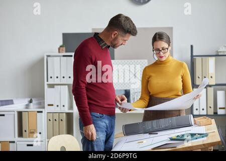 Ritratto di due architetti maturi, uomo e donna, che lavorano insieme su progetti mentre si levano in piedi da una scrivania in ufficio, copia spazio Foto Stock