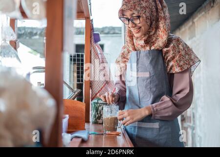 Venditore asiatico di angkringan femminile con hijab e grembiule in possesso di un bicchiere da bere in piedi sul lato del carrello Foto Stock