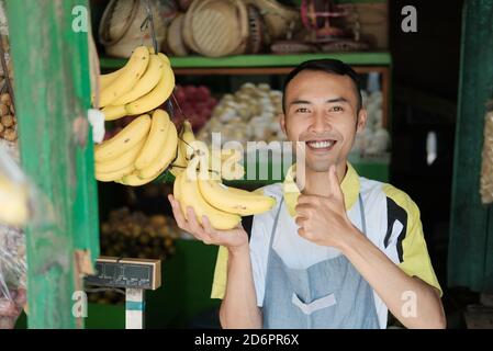 giovane uomo sorridente con banane di frutta fresca nel negozio di frutta con i pollici verso l'alto Foto Stock