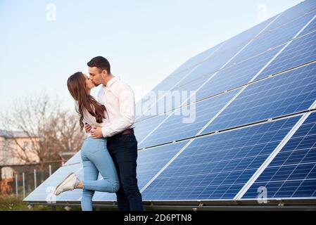 Vista laterale di una bella coppia baciante in piedi vicino a un grande modulo pv in zona rurale, primo piano Foto Stock