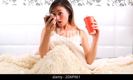 Bella giovane donna si sveglia a letto con una tazza di caffè e una sveglia. La donna ha dormito troppo ed è in ritardo, reagendo con orrore al momento. Foto Stock