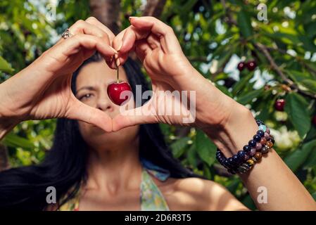 Bella giovane donna che tiene la ciliegia matura rossa e mostra la forma del cuore Con le mani fuoco selettivo Foto Stock